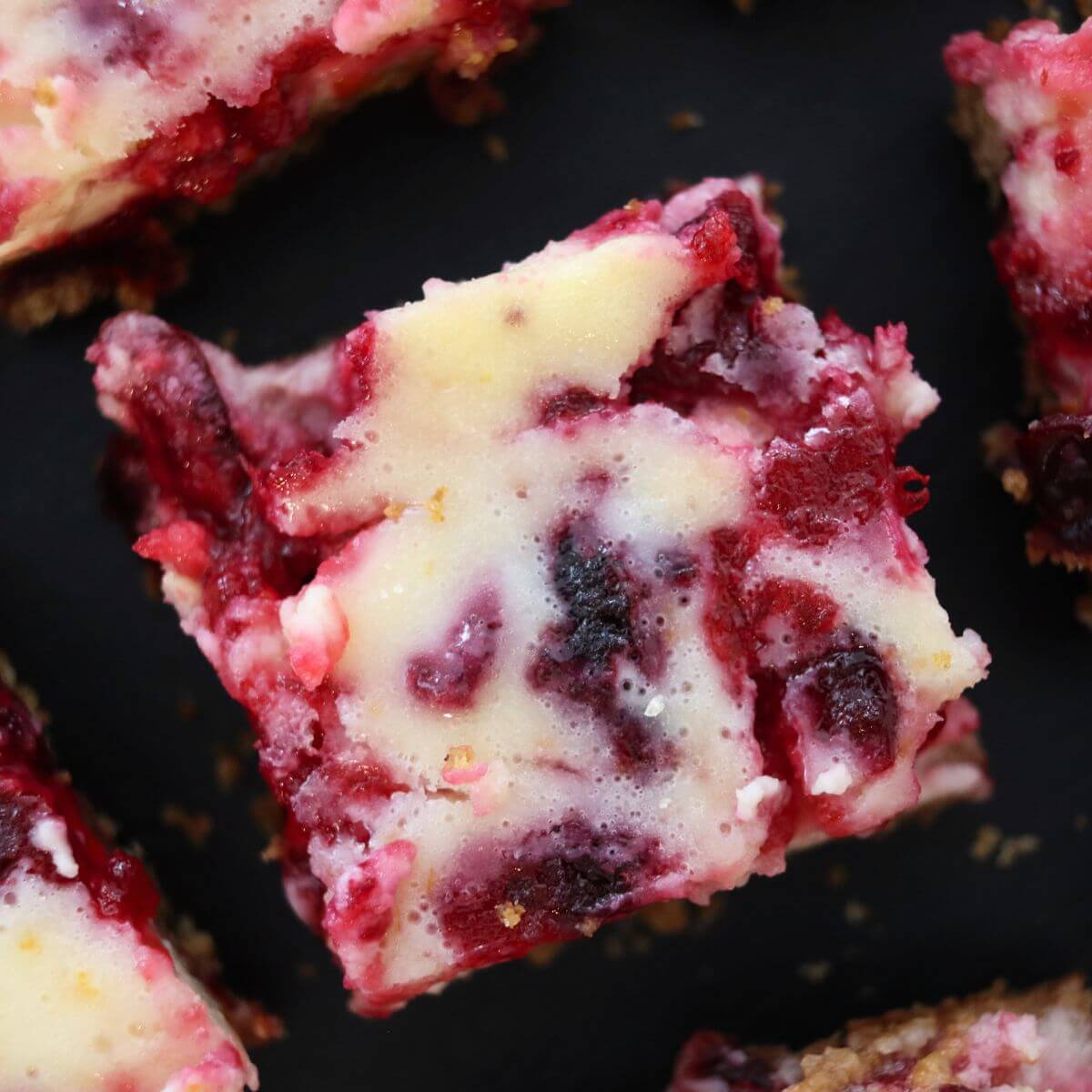 Cranberry cheesecake bars arranged on a dark surface, showing marbled cranberry topping and graham cracker crust.