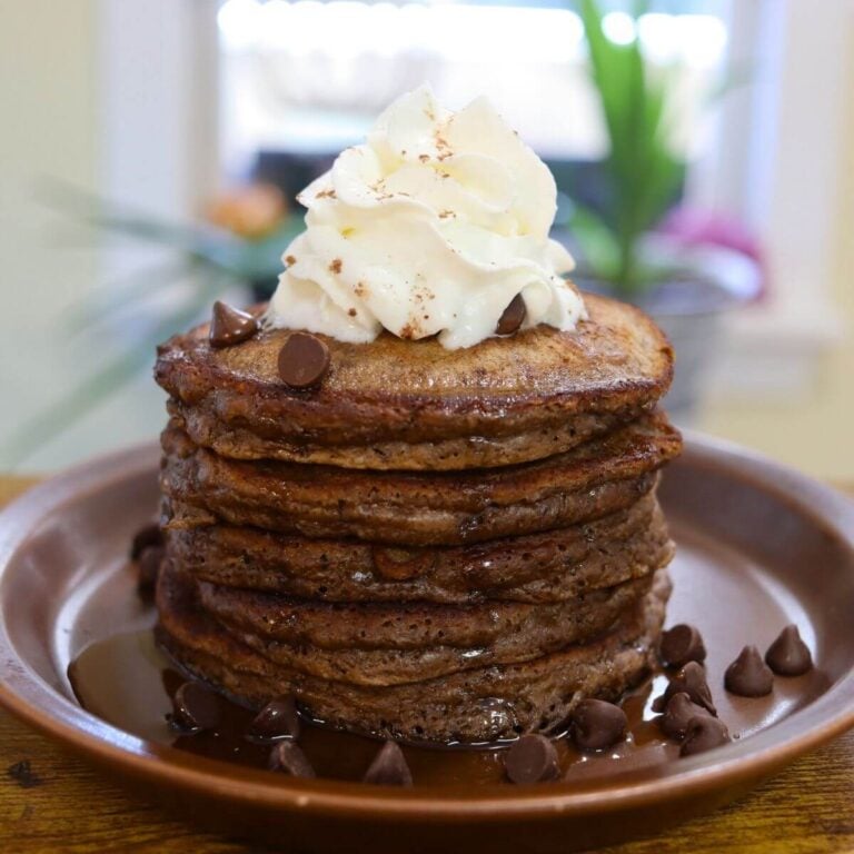 Double chocolate pancakes topped with whipped cream and milk chocolate chips on a brown plate.