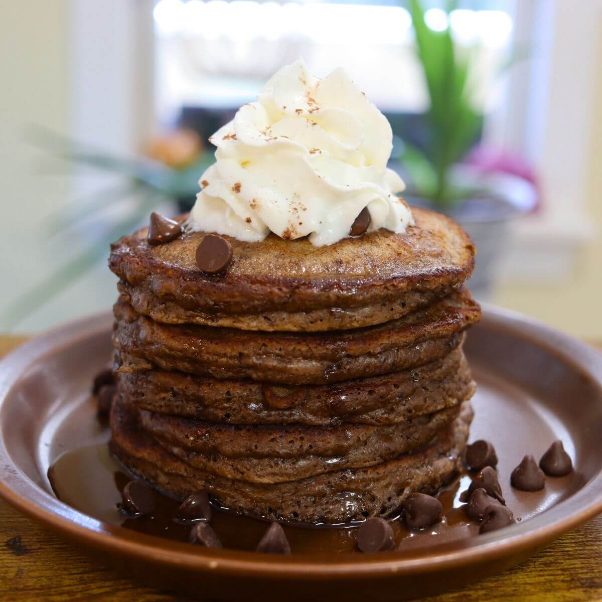 Double chocolate pancakes topped with whipped cream and milk chocolate chips on a brown plate.
